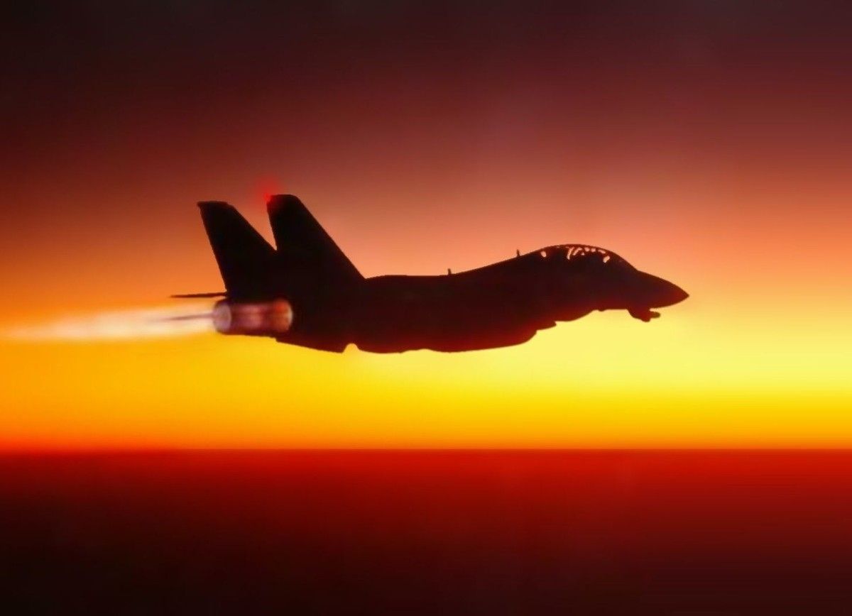 A twin-tail fighter jet silhouetted against a sunset, afterburner lit